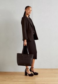 Sort totebag med to håndtag, rektangulær form, glat tekstur, med mærke på fronten. Model i en sort blazer og kjole, stående på trægulv.
