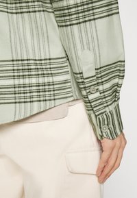 Camisa de cuadros verdes con puños abotonados, que presenta una textura ligera. Capas sobre una prenda beige con un detalle de bolsillo.