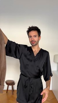 Robe en satin noir avec des manches courtes et larges et une taille liées, tenue à côté d'une veste en daim marron ; des meubles en bois visibles à l'arrière-plan.