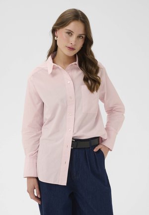 Femme aux longs cheveux bruns portant une chemise rose clair à boutons, un pantalon taille haute bleu foncé et des boucles d'oreilles en perles, debout avec une main dans la poche.