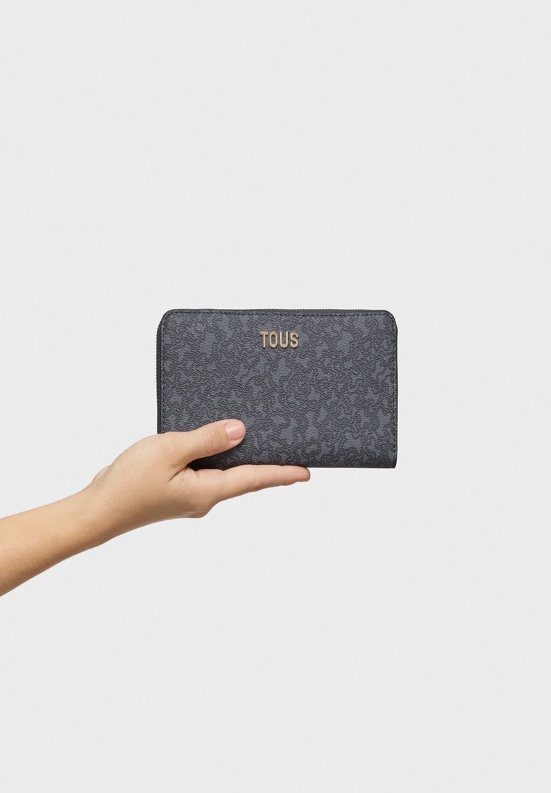 Cartera negra texturizada con un patrón sutil, logotipo dorado "TOUS", cierre de cremallera y forma rectangular, sostenida en una mano.