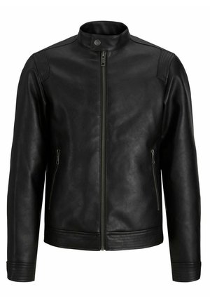 Personne portant une veste en cuir noire, une chemise marron, un pantalon beige et des baskets noir et blanc, debout devant un fond clair uni.