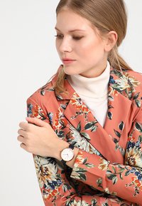 Blazer orange à motifs floraux, superposé à un col roulé crème, avec une montre or et marine à cadran rond et au design minimaliste.