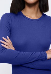 Vrouw met gekruiste armen, gekleed in een aansluitende, langjarige, geribbelde donkerblauwe top, weergegeven van nek tot taille tegen een eentonige lichte achtergrond.