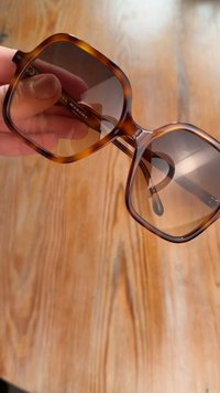 Mano che tiene occhiali da sole oversize quadrati con montatura tartarugata e lenti sfumate marroni contro uno sfondo in legno.