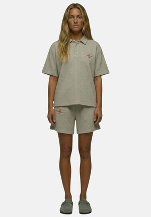 Jeune femme aux longs cheveux debout, portant un polo à manches courtes texturé gris clair et un short assorti avec des poches ainsi que des chaussures grises sans lacets.