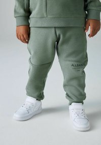 Pantalones de chándal verdes con tejido texturizado, puños acanalados y detalles en los paneles laterales. Combinados con zapatillas blancas con correas ajustables.