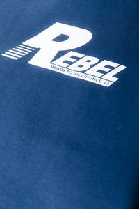 Sfondo in tessuto blu navy con testo bianco prominente che legge "REBEL" e "PRONTO DA INDOSSARE DAL '13." Texture morbida e liscia.
