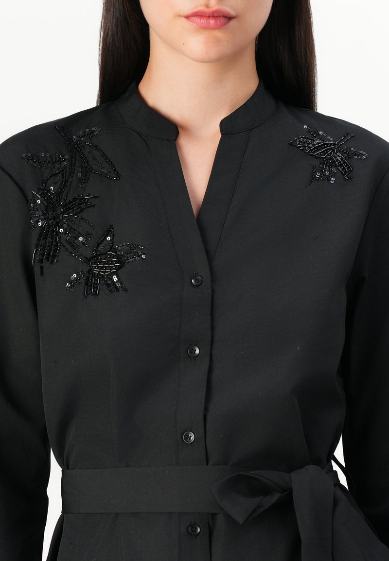 Schwarze Bluse mit Stehkragen, versehen mit filigranen Perlen- und Paillettendekorationen in Blumenform auf einer Schulter und einem gebundenen Taillenband.