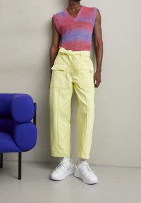 Persona con una camiseta sin mangas de tonos rosas y morados a rayas, pantalones amarillos de cintura alta con cinturón y zapatillas blancas, de pie al lado de una silla azul.