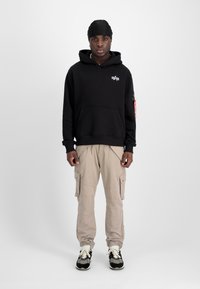 Schwarzer Kapuzenpullover mit Fronttasche und Logo, beige Cargo-Shorts mit Seitentaschen und schwarze Sneakers mit weißen Schnürsenkeln.