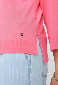 Maglione rosa chiaro realizzato in morbido tessuto a maglia, con orlo a costine, spacco laterale e un piccolo dettaglio del logo metallico in basso a sinistra.
