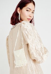 Perleveske i hvite perler, med en teksturert overflate og frynset bunn, brukt sammen med et rynket, beige plagg.