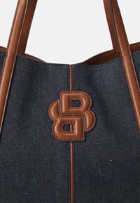 Denim-kangaskassi, jossa on ruskeat nahkakoristelut ja -olkaimet. Etupuolella on huomattava ruskea nahkalogo.
