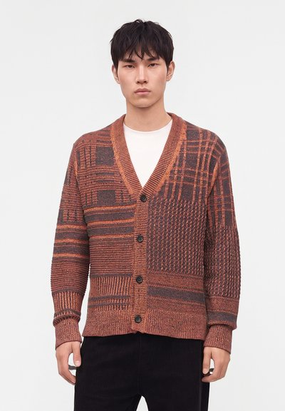 Homme portant un cardigan boutonné à motifs rouille et gris foncé par-dessus une chemise blanche et un pantalon noir sur un fond clair uni.