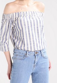 Blouse rayée bleu et crème, décolletée aux épaules, avec un col froncé et des manches trois-quarts, associée à un jean bleu clair.