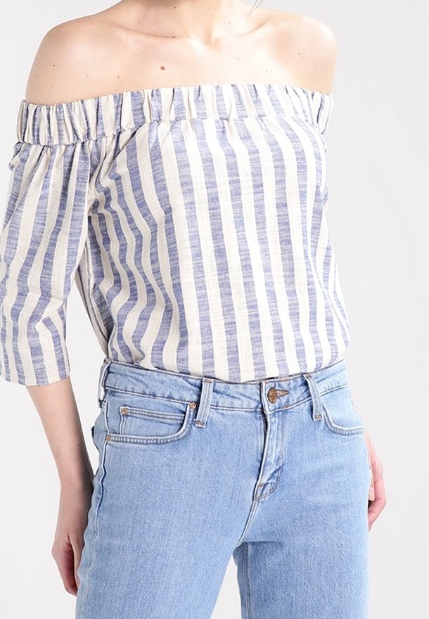 Blouse rayée bleu et crème, décolletée aux épaules, avec un col froncé et des manches trois-quarts, associée à un jean bleu clair.