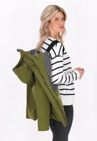 Olivgrön jacka med grå insida, fullt dragkedja och tryckknappar, bärs över en vit randig långärmad skjorta, modellen poserar i profil.