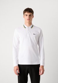 Langermet hvit polo-skjorte med svart og gulstripet krage. Har en liten svart "BOSS"-logo på brystet. Myk bomullsmateriale.