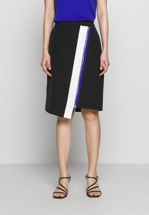 Vrouw draagt een zwarte knielange rok met een verticale witte en blauwe ritssluiting, gecombineerd met zwarte sandalen met bandjes en een blauwe top.