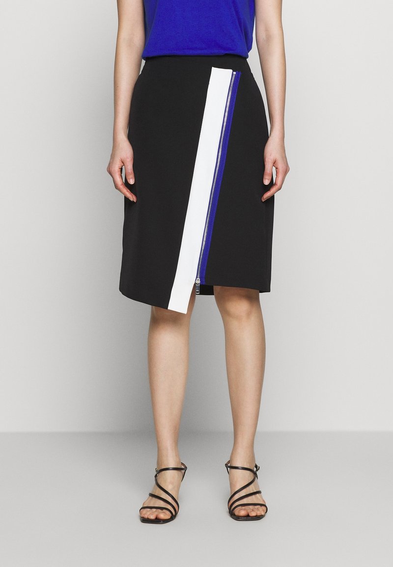 Femme portant une jupe noire mi-longue avec un détail de fermeture éclair verticale blanche et bleue, associée à des sandales noires à lanières et un haut bleu.