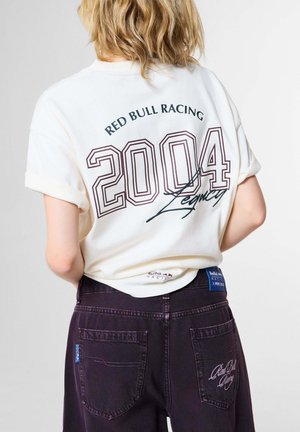 Person mit blonden Haaren trägt ein weißes Red Bull Racing Shirt mit dem Text "2004 Legacy" und dunkle Hose mit Red Bull Racing Stickerei auf den hinteren Taschen.