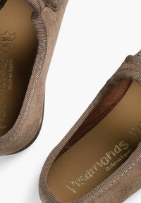 Zapato de ante marrón sin cordones con parte superior texturizada y forro interior en contraste. Logo dorado grabado en la plantilla. Diseño de suela plana.