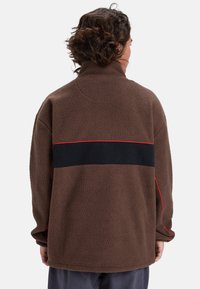 Chaquetón de forro polar marrón con un alto cuello, que presenta un panel negro y una franja de acento rojo a lo largo de la espalda. Textura suave; corte relajado.