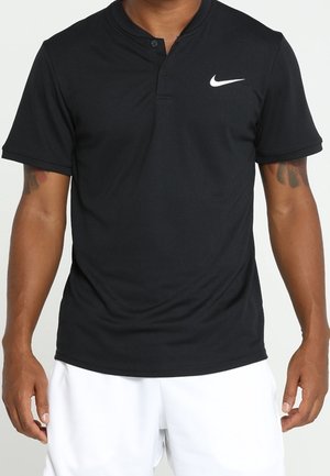 Man draagt een zwart Nike-poloshirt met korte mouwen, voorzien van twee knopen, en witte korte broek, staand met ontspannen armen langs het lijf.