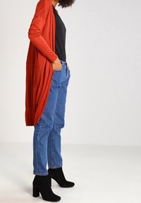 Langer, offener Cardigan in lebhaftem Rot, kombiniert mit einem schwarzen Shirt und blauen Jeans. Das Model trägt schwarze, hohe Stiefeletten. Glattes Stoffegefühl.