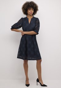 Tengerészkék csipkeborítású ruha puffos ujjal és V-nyakkivágással. Virágmintákkal és bővülő sziluettel rendelkezik; fekete magassarkúval párosítva.