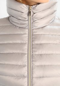 Jasnoszara kamizelka puchowa z wysokim kołnierzem; wzór pikowany, błyszcząca tekstura; posiada centralny zamek błyskawiczny z elementami w kolorze złotym.