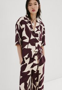 Kvinde iført matchende løs kortærmet skjorte og bukser med abstrakt mønster af mørklilla og cremefarvede blade, står med hænderne i lommerne.