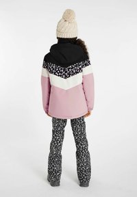 Giacca da sci rosa e nera con sezioni bianche e decorate con motivi floreali. Abbinata a pantaloni neri con motivo. Berretto in maglia beige con pom-pom.