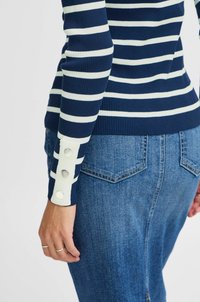 Haut en tricot rayé bleu marine et blanc avec une texture côtelée, présentant de gros boutons aux poignets. Associé à un jean en denim bleu clair.