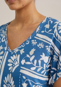 Zilas kokvilnas topiņš ar V-veida kakla izgriezumu, dekorēts ar baltām ziedu un lineārām rakstiem. Mīksta tekstūra, īsas piedurknes, radīts komfortam.