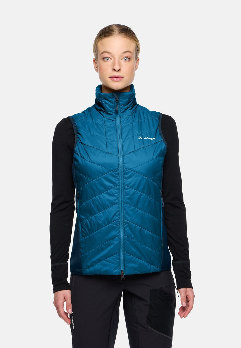 Türkiser isolierter Weste mit hohem Kragen, vertikalen Nähten, Reißverschluss vorne und Logo-Detail auf der linken Brust. Schwarzes Langarm-Basisschicht darunter.