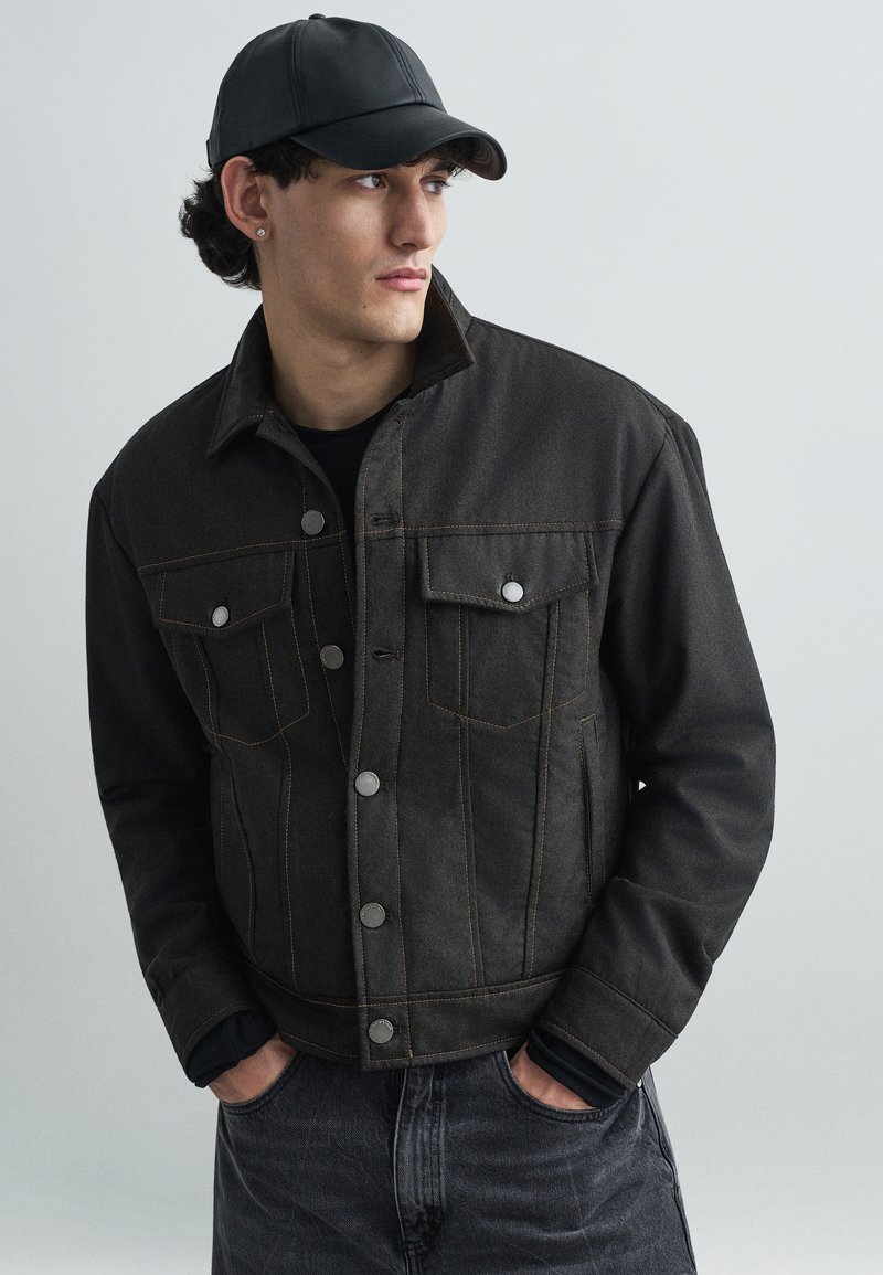 Sort denimjakke med orange syninger, trykknapper og to brystlommer. Båret over en sort skjorte og parret med mørke jeans.