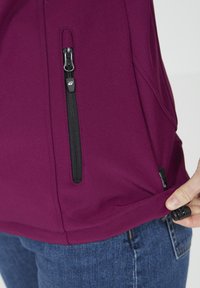 Vinröd softshell-jacka med en dragkedjeficka, svart dragkedjehuvud och dragskojustering i nederkant. Mjuk textur, figursydd design.