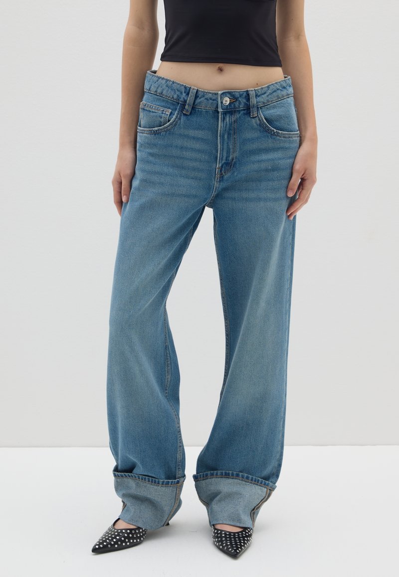 Helle blaue Schlagjeans aus weichem Denim mit fünf Taschen, einem Knopfverschluss und umgeschlagenen Säumen. Kombiniert mit schwarzen spitz zulaufenden Schuhen mit weißen Punkten.