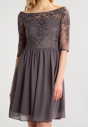 Femme portant une robe gris foncé mi-longue, bustier décolleté avec broderies florales en dentelle et manches trois-quarts transparentes.