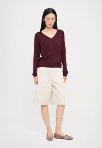 Burgunderfarbener V-Ausschnitt-Pullover, cremefarbene Shorts und gemusterte Ballerinas. Weicher Stoff, lockere Passform und Ärmel in Ellenbogenlänge. Schlichtes Design ohne Verzierungen.