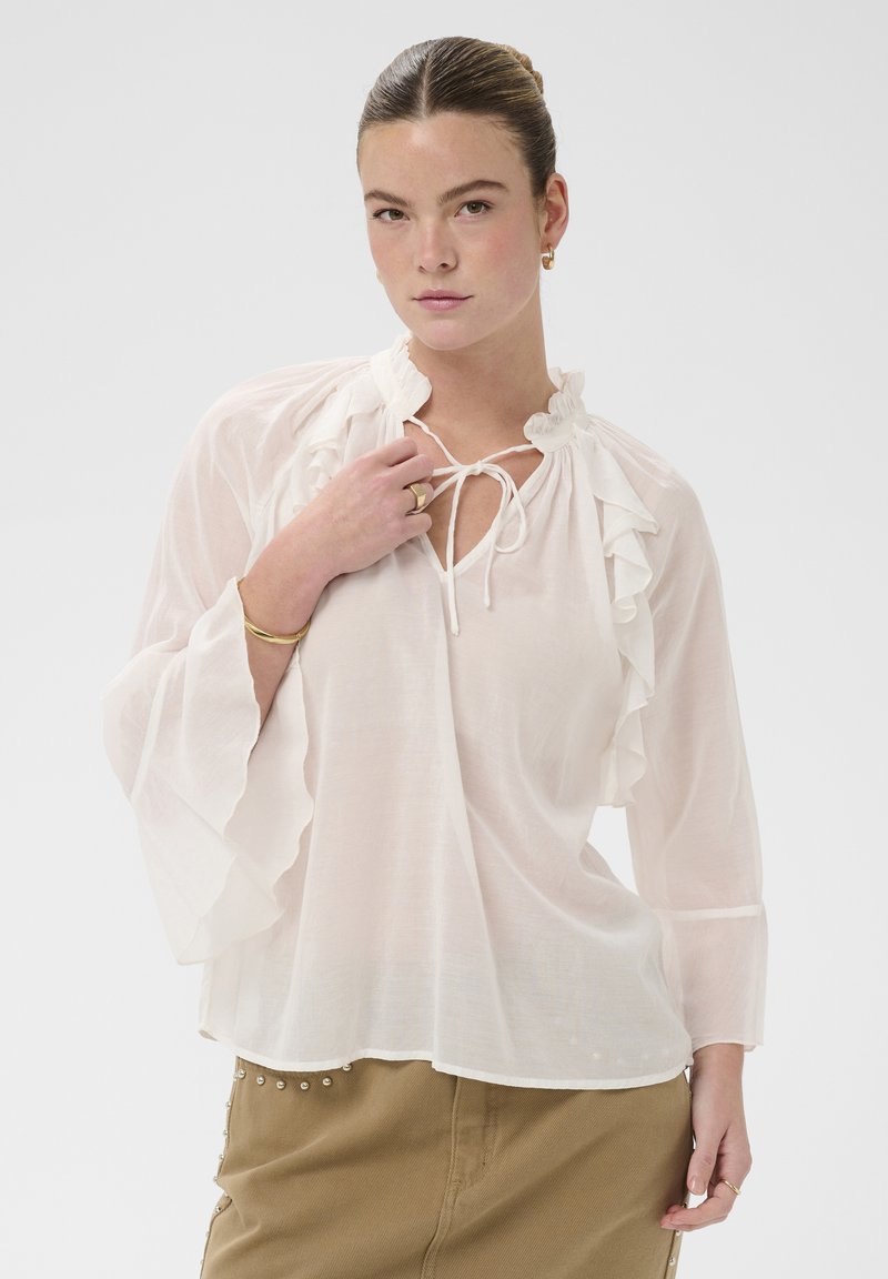 Jeune femme portant un chemisier blanc transparent avec un col et des manches à volants, une jupe beige ornée de détails en perles, et des bijoux en or, regardant devant elle.