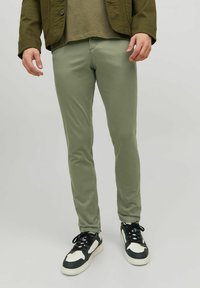 Chinos slim-fit vert olive en tissu lisse, avec cinq poches et une fermeture à bouton, associés à des baskets noires et blanches.
