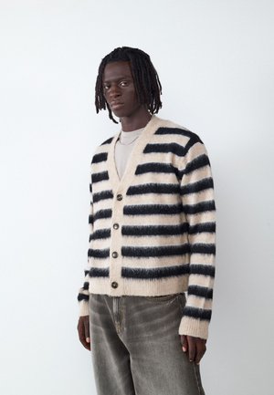 Joven con dreadlocks cortos lleva un cárdigan borroso beige y negro con rayas sobre una camisa clara y vaqueros grises, de pie frente a un fondo blanco.