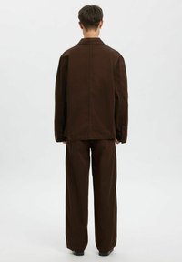 Brązowa bawełniana kurtka z dopasowanymi szerokimi spodniami o prostych nogawkach. Posiada klasyczny kołnierz i mankiety na guziki, cechuje się czystym, minimalistycznym designem.