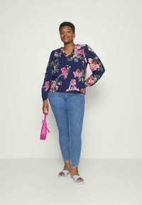 Blusa azul-marinho com padrão floral, mangas compridas e decote em V; combinada com jeans skinny azul-claro e sandálias cinzentas; a usar uma bolsa rosa.