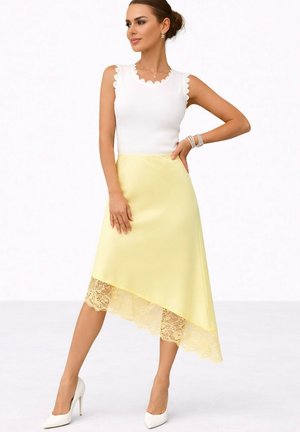 Femme posant en haut blanc sans manches et jupe asymétrique jaune pâle avec ourlet en dentelle, portant des talons blancs et des bijoux.