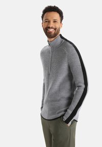 Grauer Strickpullover mit einem Viertelreißverschluss und schwarzen Seitenstreifen; besitzt eine gerippte Textur und eine figurbetonte Passform. Gekleidet mit olivgrünen Hosen.