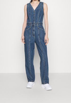 Combinaison en jean bleu sans manches avec boutons à l'avant et poches, portée avec des baskets blanches épaisses sur une personne debout.